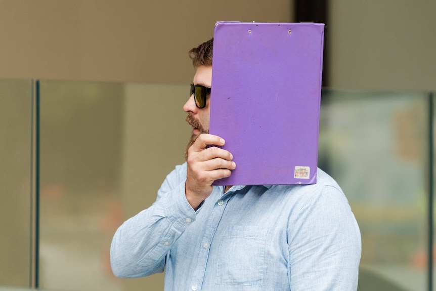 A mid-shot of a young man with a beard and sunglasses shielding his face with a purple clipboard outside court.