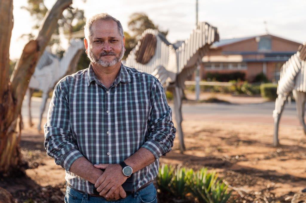 Peter wearing a blue check shirt, standing outside in the rural town of Norseman, with gumtrees and dirt.
