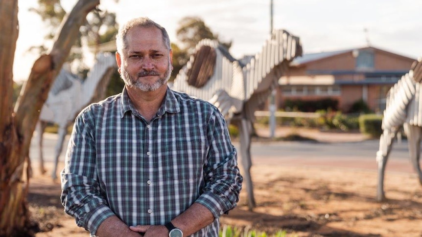 Peter wearing a blue check shirt, standing outside in the rural town of Norseman, with gumtrees and dirt.