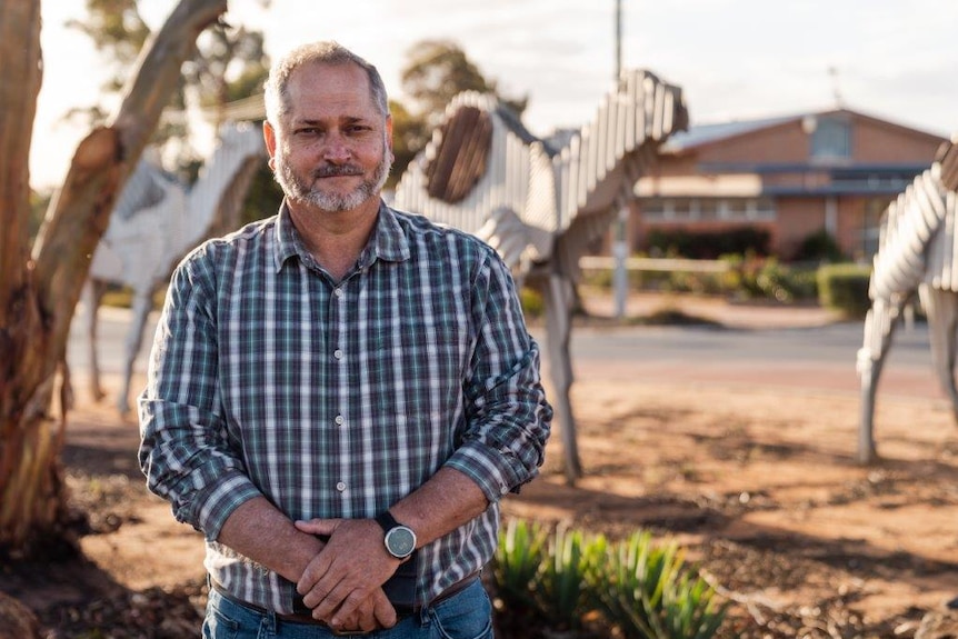 Peter wearing a blue check shirt, standing outside in the rural town of Norseman, with gumtrees and dirt.