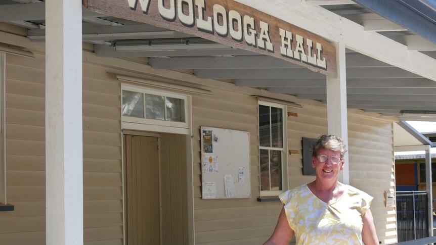 A lady stands smiling in the sun in front of a timber building with the sign 'Woolooga Hall'.