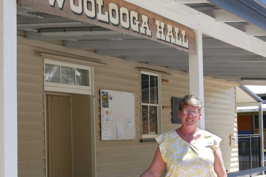 A lady stands smiling in the sun in front of a timber building with the sign 'Woolooga Hall'.