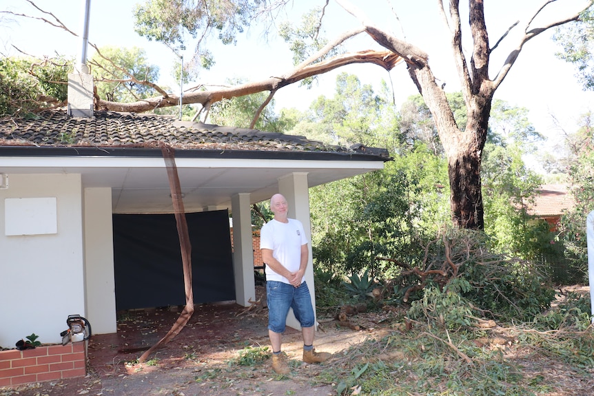 A man stands at the front of his house with a large tree branch lying on the roof.