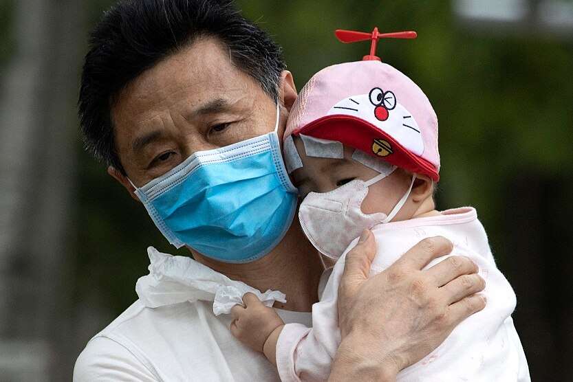 A man wearing a face mask holds a baby who is also wearing a mask and grasping a tissue.