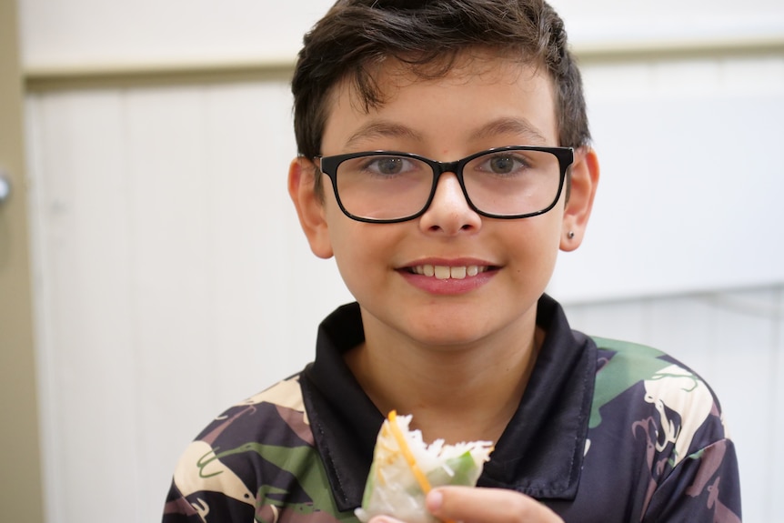 A boy holds a half eaten rice paper roll and smiles.