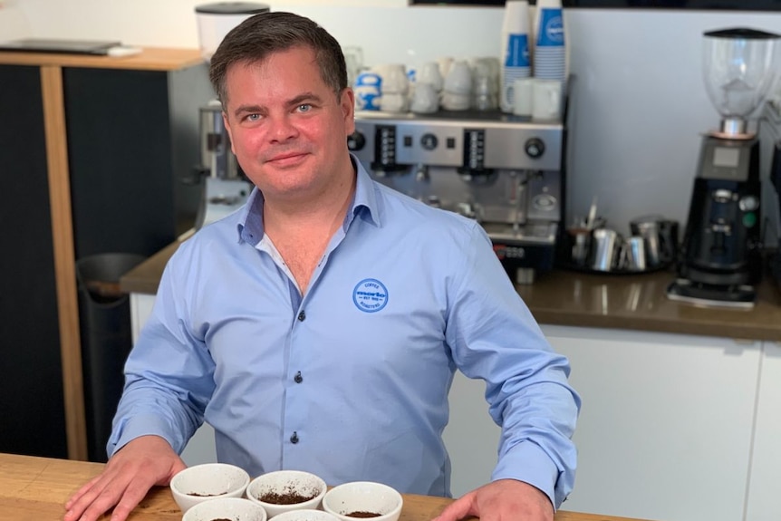 A man in a blue collared shirt stands at a counter with several cups of ground coffee.