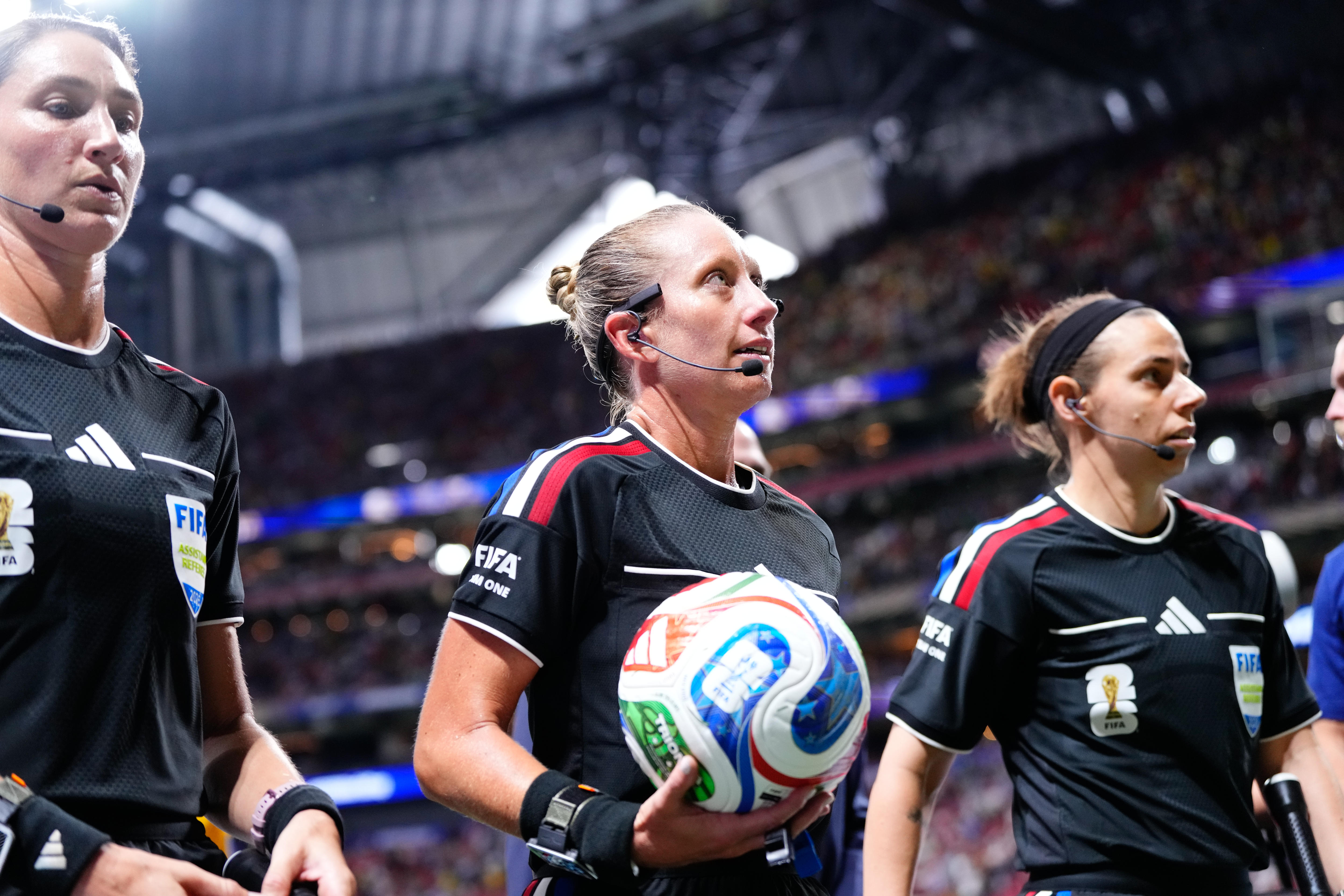 Three female soccer referees in black walk out in a stadium, with the one in the middle holding a ball