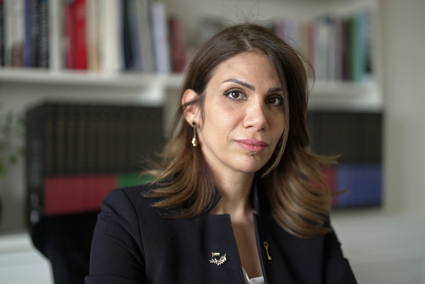 A woman sitting in a room, looking directly at the camera. Behind her is a bookshelf.