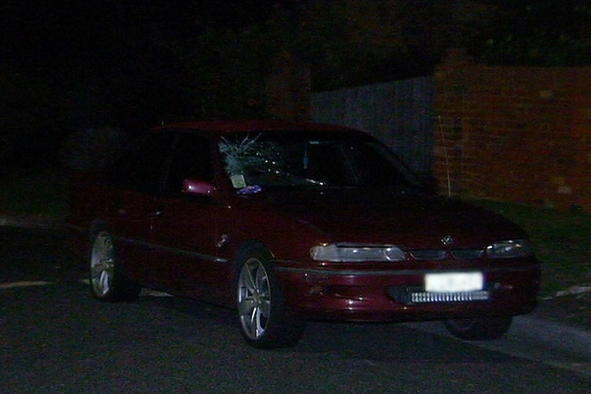 A red sedan parked on a street in the dark with a smashed windshield.