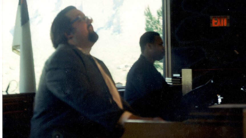 Two male musicians sit side by side; the man in the foreground plays an organ.