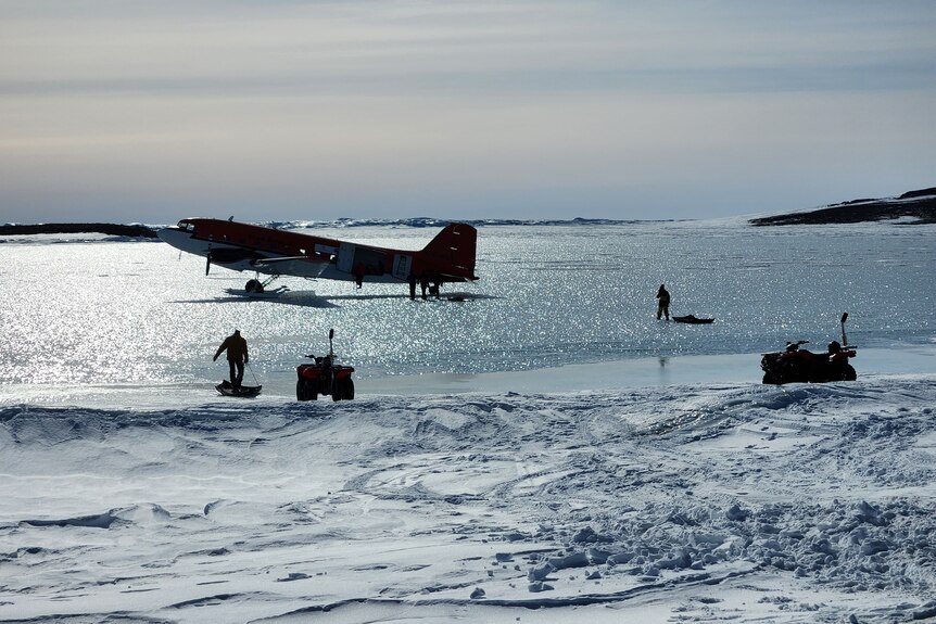 A propeller plane sits on shimmering ice, as a man carries cargo over to it.