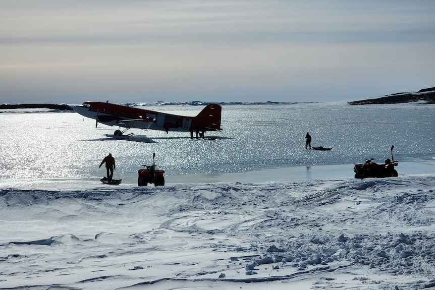A propeller plane sits on shimmering ice, as a man carries cargo over to it.