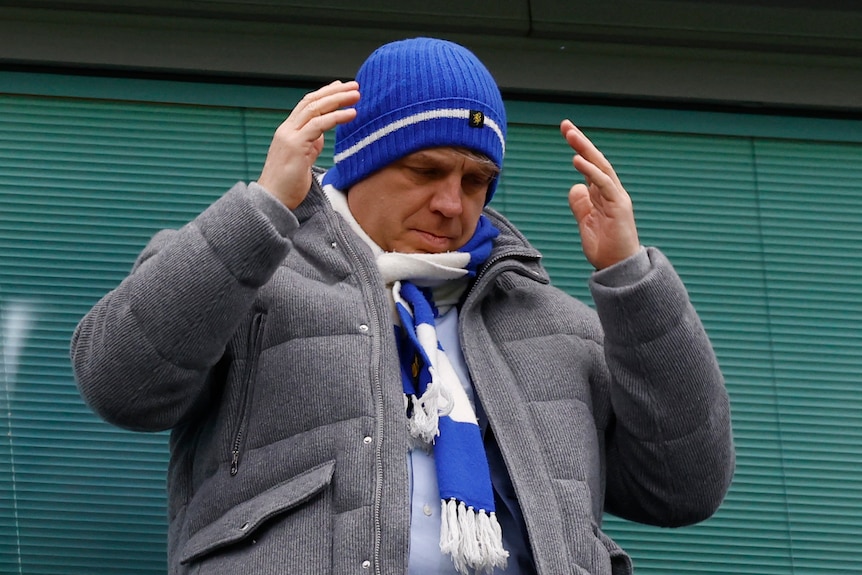 A man wearing a blue beanie and a blue and white scarf gestures with his hands.