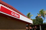 A red Australia Post sign against a blue sky with trees in the background.