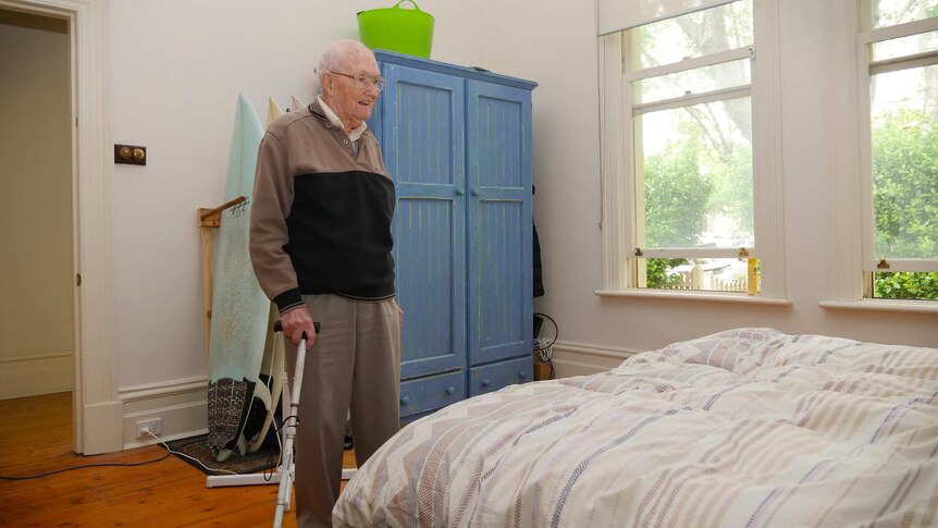 Stan McKay smiles while standing in a light-filled room with a double bed and a blue wardrobe.