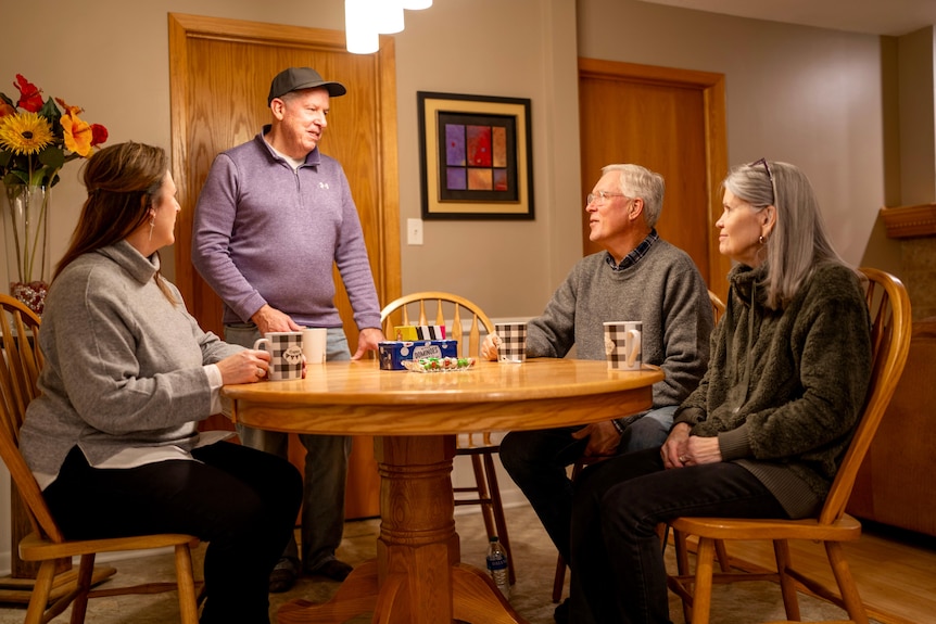 The friends sit around a dining room table and chat. There are coffe mugs on the table. Scott stands in front of a door.
