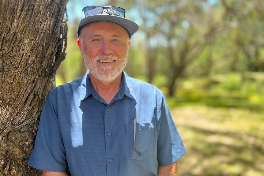 A man in a blue shirt smiles, standing next to a tree.