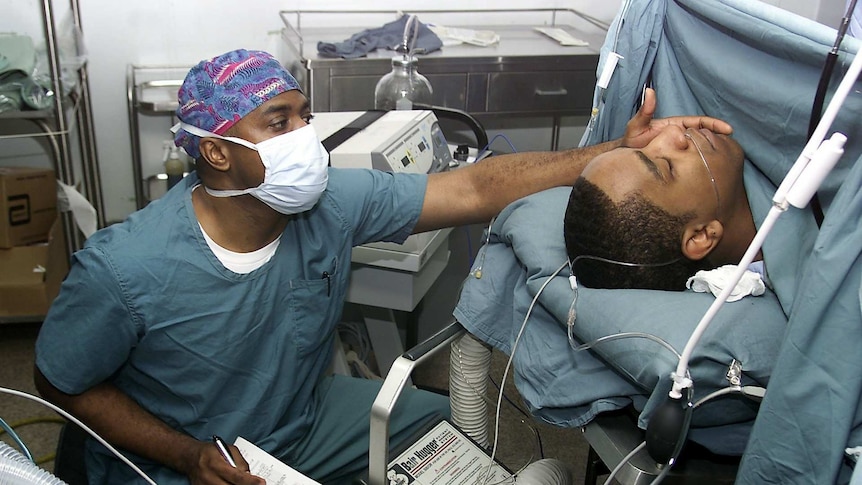 Two men in an operating theatre, one under anaesthetic lying down and the other monitoring him.