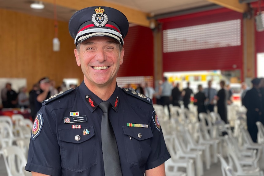Middle aged man smiling in QFES uniform, dark grey tie, dark blue shirt, cap, medals, rows of seats behind.
