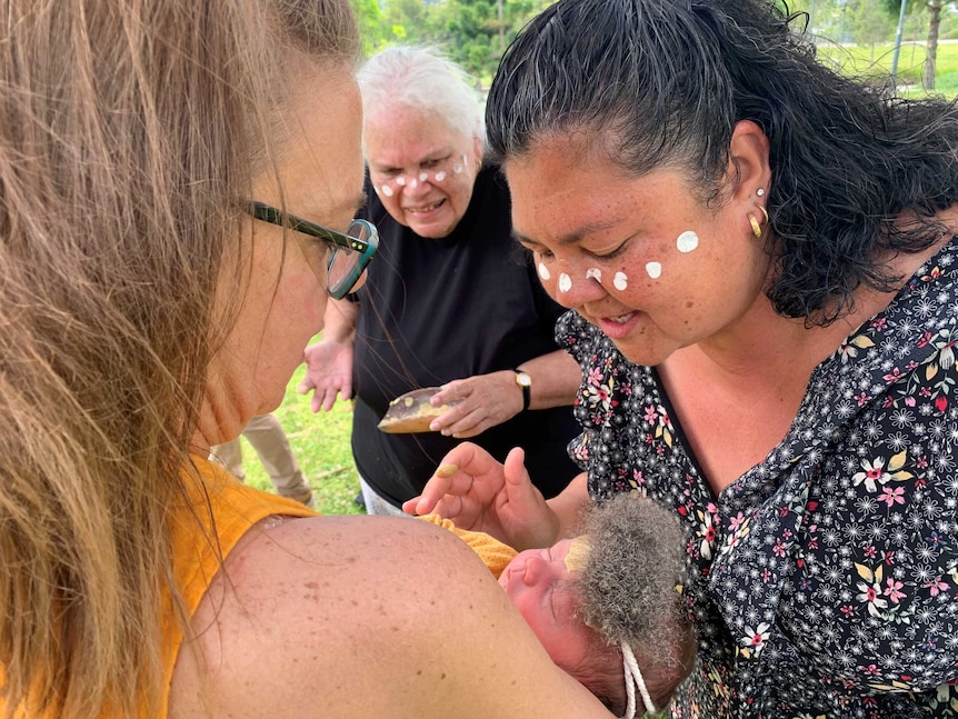 A woman holds a baby while another woman paints its face with ochre