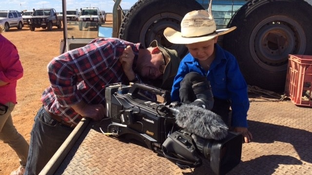 Cameraman Campbell Miller shows a young boy wearing a cowboy hat sitting on the back of a ute his TV camera.