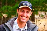 A widely smiling man in a cap, sunglasses on top, blue jacket, light blue shirt, in front of trees.