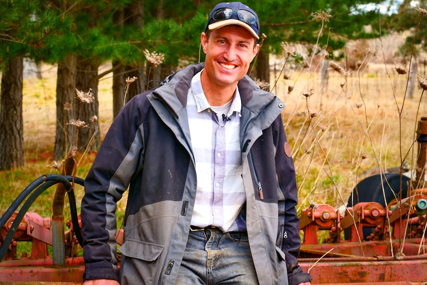 A widely smiling man in a cap, sunglasses on top, blue jacket, light blue shirt, in front of trees.