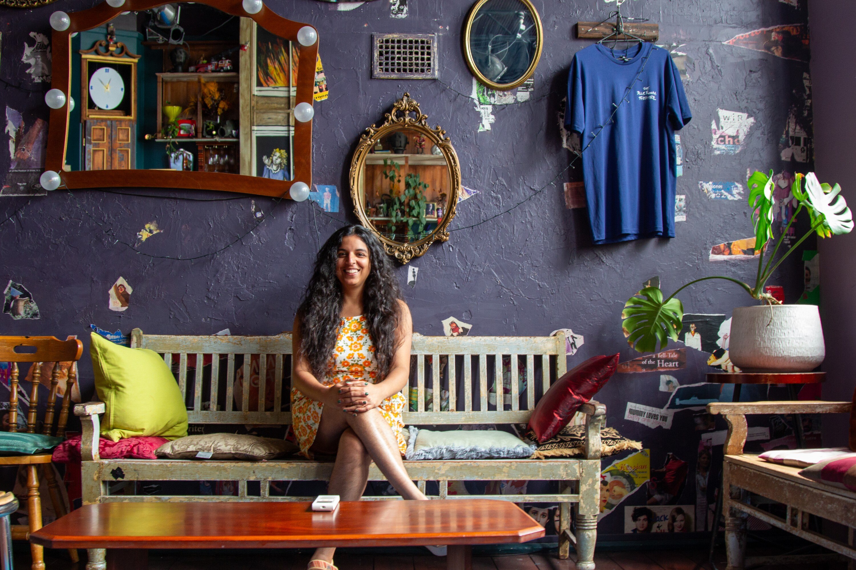 Uma mulher australiana do sul da Ásia, na casa dos 30 anos, está sentada, sorrindo, com as pernas cruzadas, no saguão azul decorado com espelhos de um teatro.