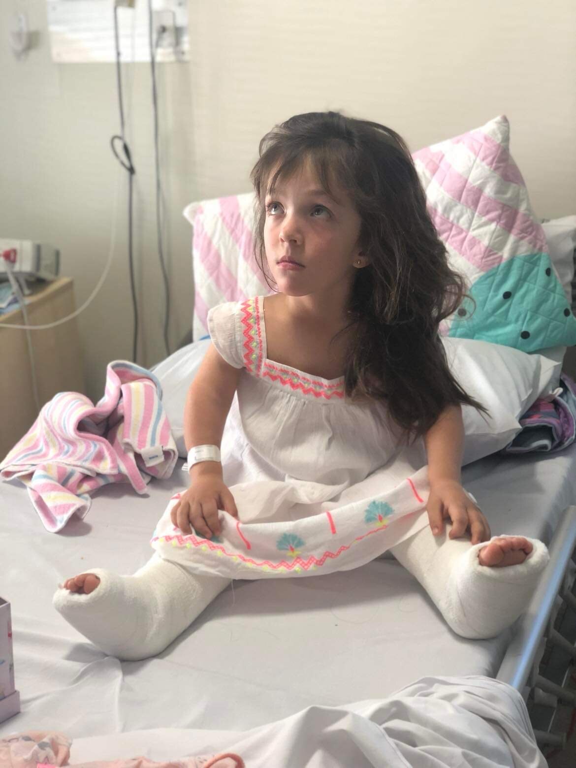 a little girl with dwarfism sits on a hospital bed, both her legs are wrapped in casts and she's wearing a pink and white dress