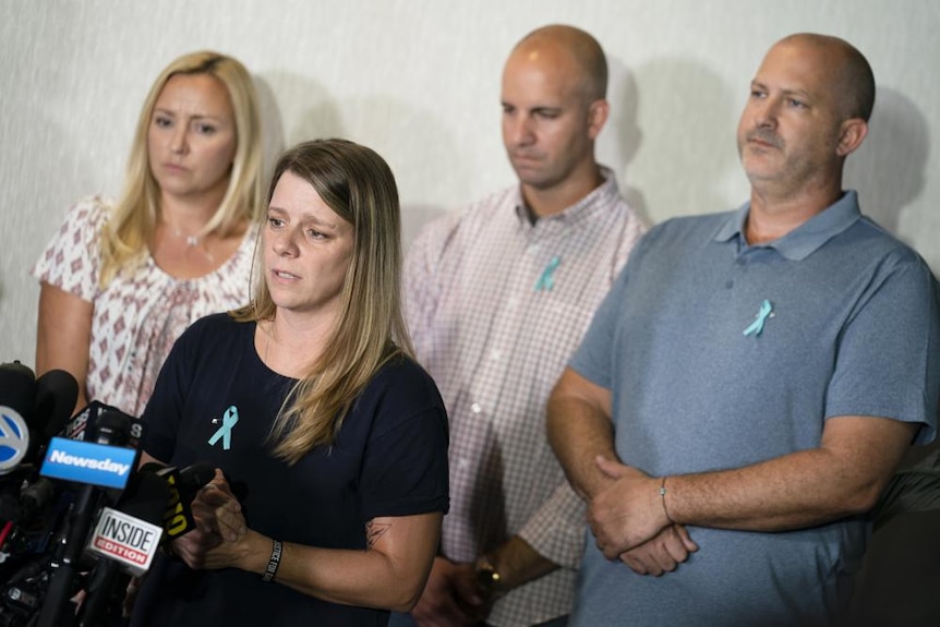 A group of men and women stand at a lectern with solemn facial expressions making a speech to media