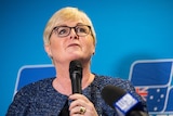 Linda Reynolds stands at a lectern with a microphone against a blue background.