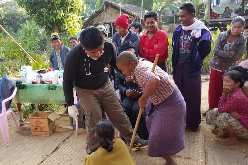 A man with a stethoscope around his neck helps an elderly person on crutches