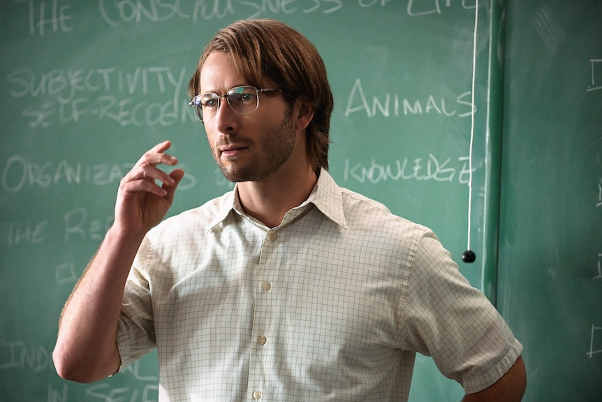 A blond man in a short sleeve button down and glasses in front of a chalkboard