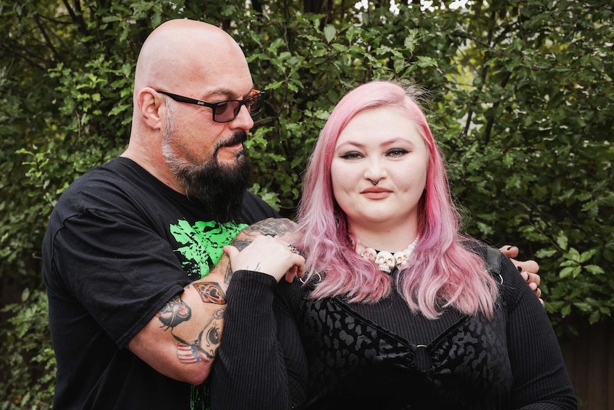 A man wearing glasses and a black shirt with his arms on the shoulders of a woman with pink hair in a garden.
