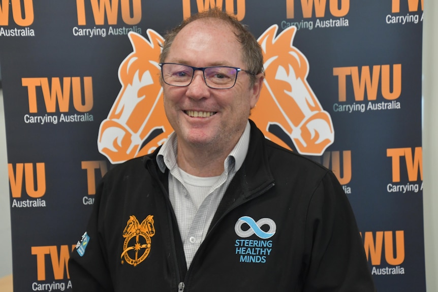 A man wearing black reading glasses and a black shirt with colourful logos stands in front of a backdrop with TWU. 