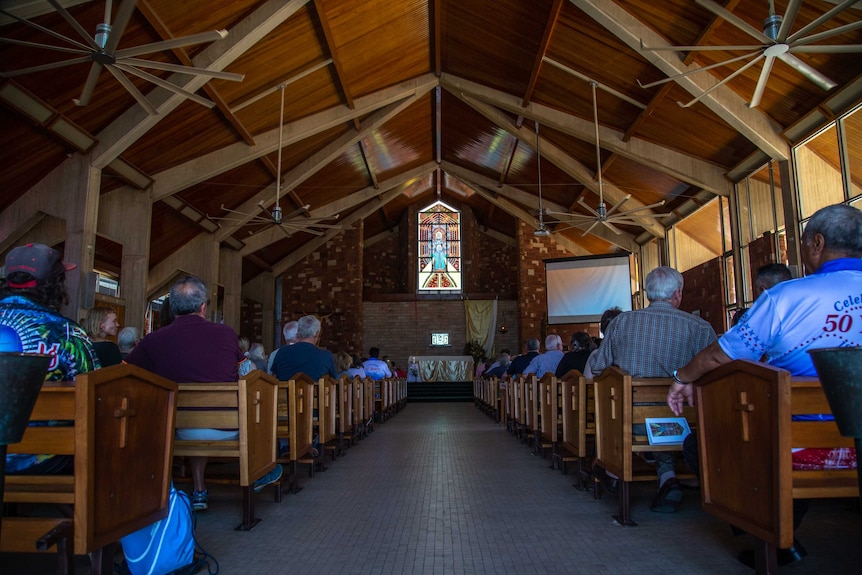 The inside of a church with parishioners in pews and a stained-glass window in the front apex.