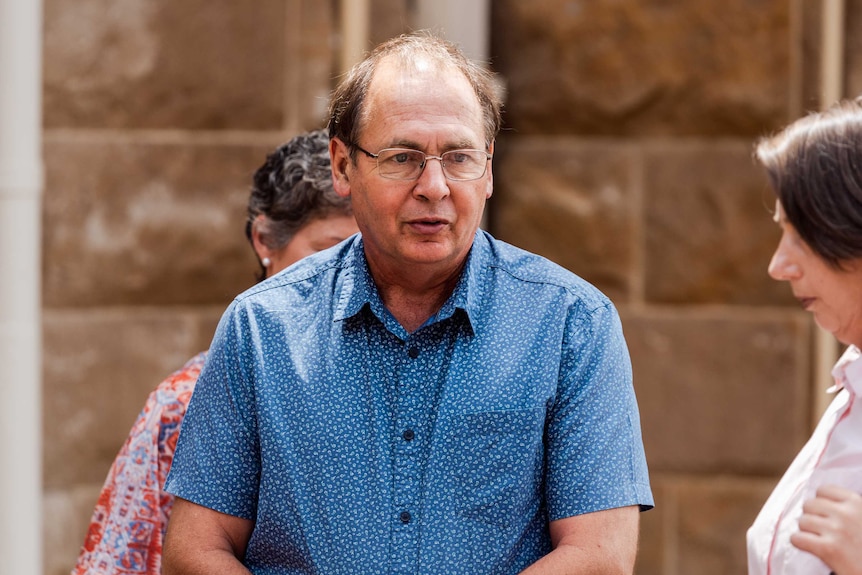 A mid-shot of a man wearing a blue shirt and spectacles with a woman behind him and another to his left.