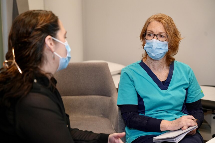 Fiona sitting in a chair across from a female patient, listening, both are wearing blue surgical masks.