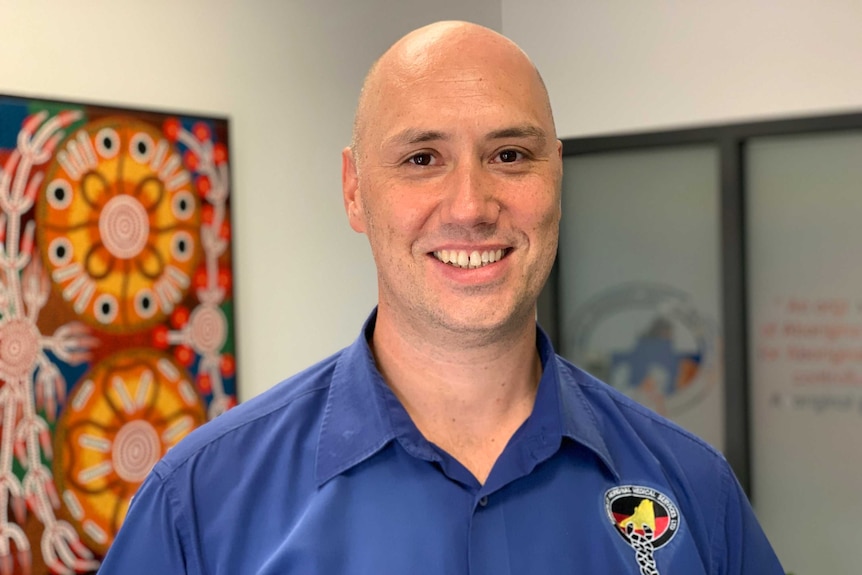 A tall man with a shaved head and blue shirt stands facing the camera in an office