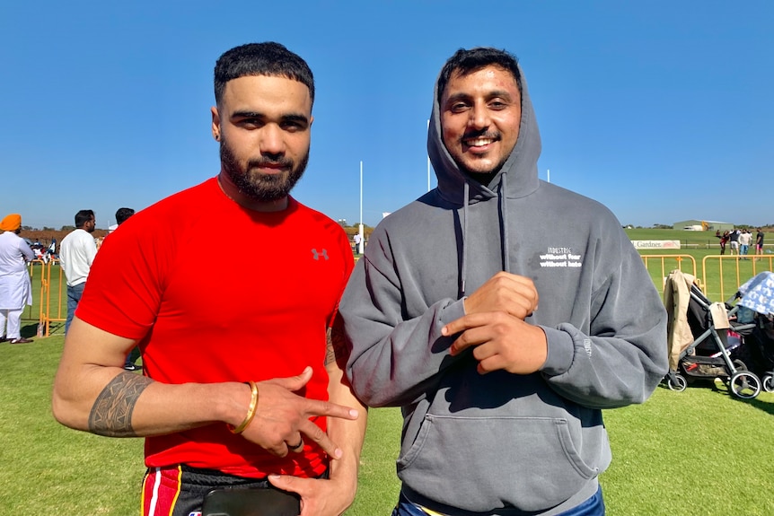 A man in a red top and a man in a grey hoody smile in front of a footy oval