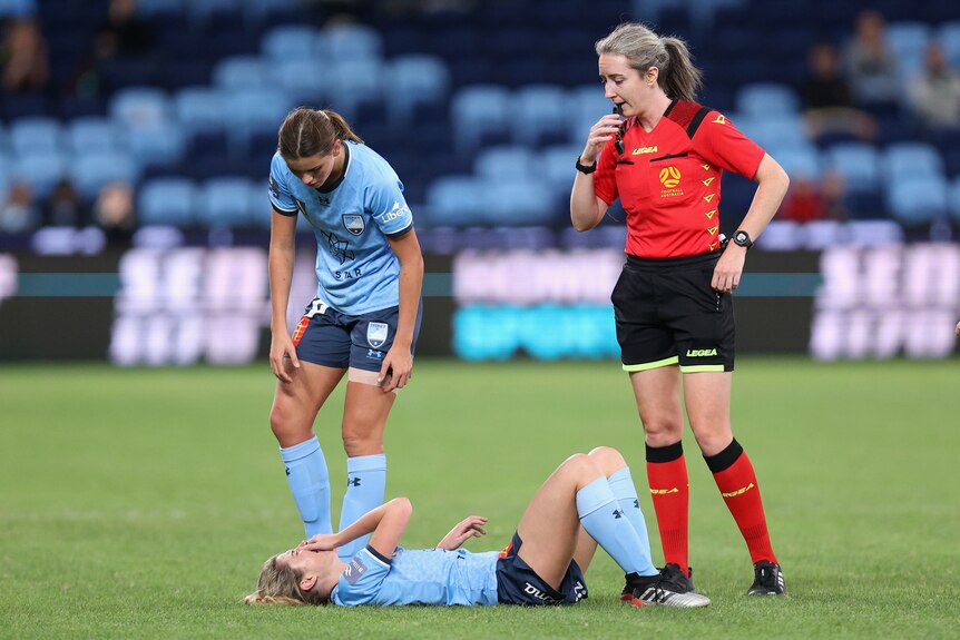 A soccer player lies on the grass holding her face as a team-mate looks on and a referee blows a whistle