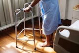 An older person in a gown walks with a walking frame with their head unseen.