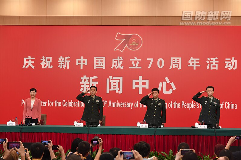 Three men wearing  Chinese military uniform stand and salute in front of a red poster celebrating 70 years of communist rule.