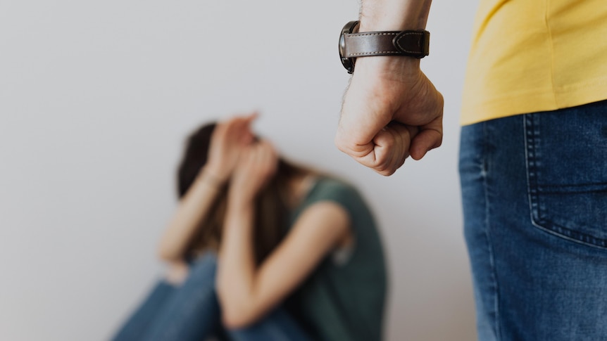 A blurred image of a woman cowering on the floor in the background, a man's clenched fist in the foreground. 