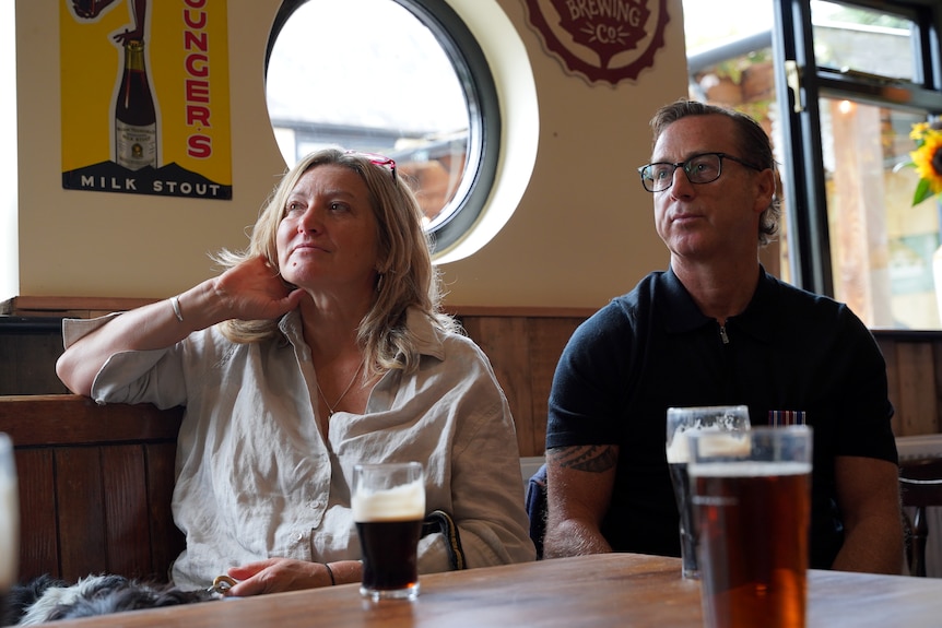 Man and woman sit in pub with beer in front of them looking off camera.
