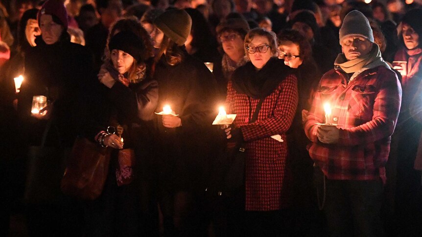 Men and women dressed in coats and beanies, hold candles in the darkness, among a large crowd.
