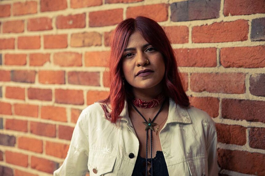 A woman in alternative country clothes leans against a brick wall.