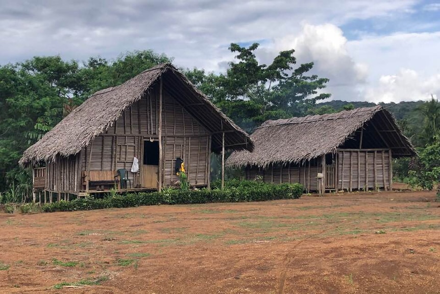 Two wooden huts stand with a field of red dirt in front and lush green trees behind