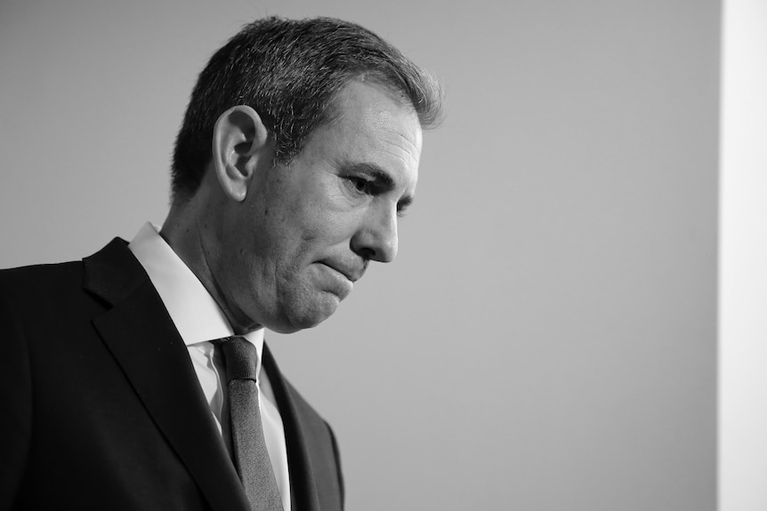 A black and white photo shows a middle-aged white man with short grey hair in a suit looking sombre.
