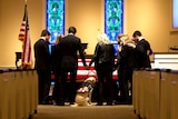 A group of people dressed in black gathered around a coffin draped in an US flag, with a yellow labrador sitting beside it.